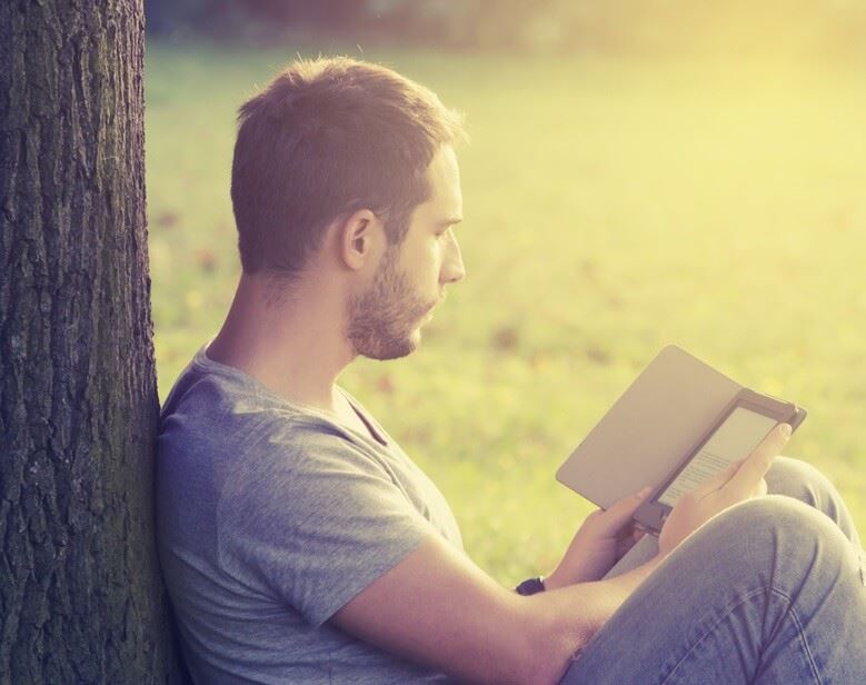 Young adult male sitting in field leaned up against a tree reading on his kindle
