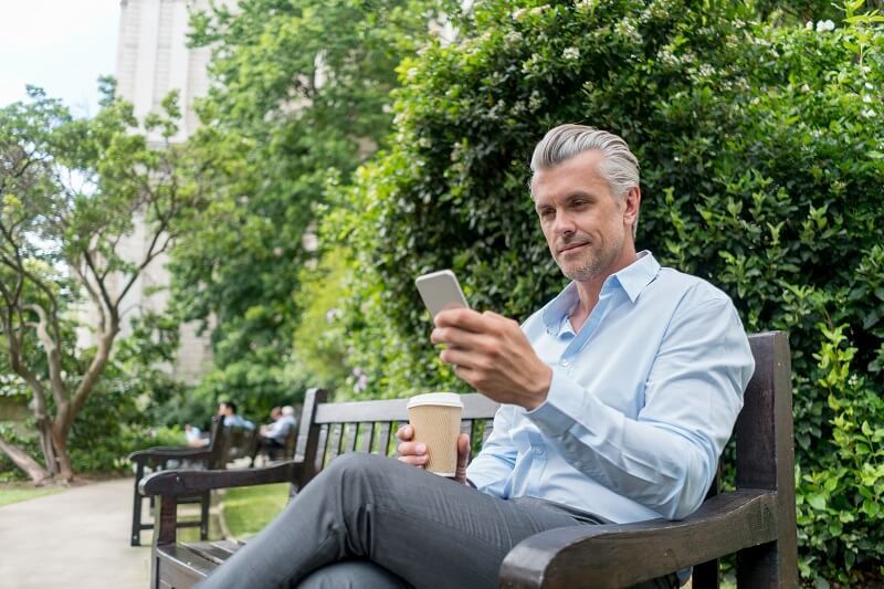 Older gentlemen sitting on a park bench with coffee in one hand while looking at his phone
