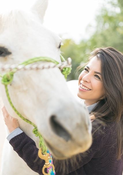 A caucasian woman with brunette hair petting a white horse