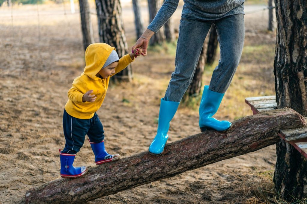 Little boy in rain boots being helped walking up a log by his parent