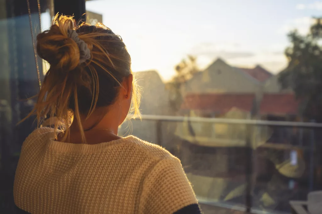 Woman looking through the window at sunset. She is alone and looks a little ad or depressed.