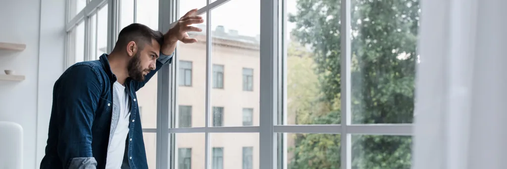 Frustration, feeling bad, self-isolation and stay home alone, deep depression, anxiety and agoraphobia. Upset attractive young bearded guy looks at large window in living room interior, panorama