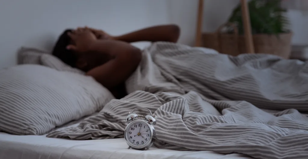 Sleepless, woman suffering from insomnia, sleep apnea or stress, headache or migraine. Tired and exhausted african american lady awake in middle of night, near alarm clock with time in dark, panorama