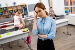 Tired teacher in blue blouse standing in front of desks and touching face