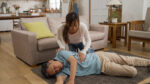 Asian adult daughter running into living room trying to wake up her unconscious father lying on floor in the living room at home